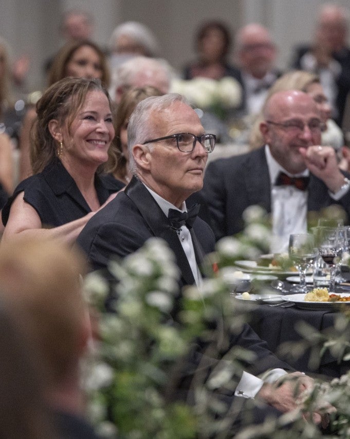 Man sitting at table, flowers in the foreground, listening to someone speaking on a stage. (Photo Credit: Wingate Downs)