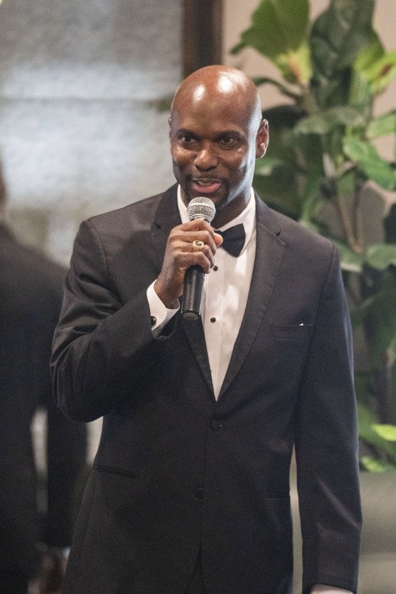 Man speaking into a microphone, dressed in a tuxedo. (Photo Credit: Wingate Downs)