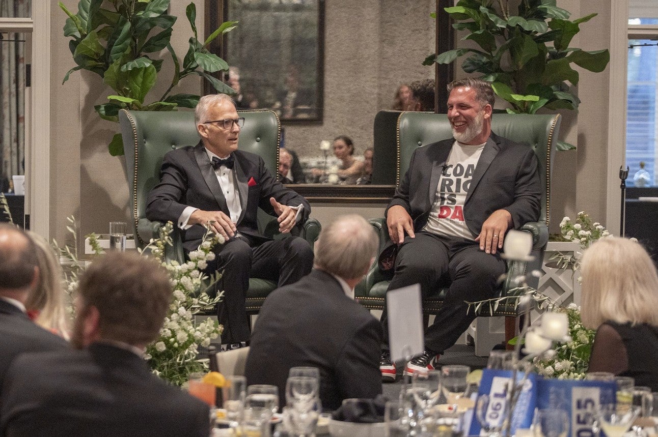 Two men sitting in chairs on a stage talking to one another. (Photo Credit: Wingate Downs)