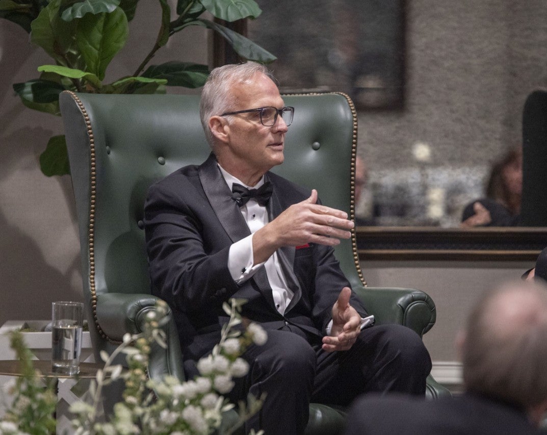 Gentleman sitting in an armchair on stage speaking to someone out of frame. (Photo Credit: Wingate Downs)