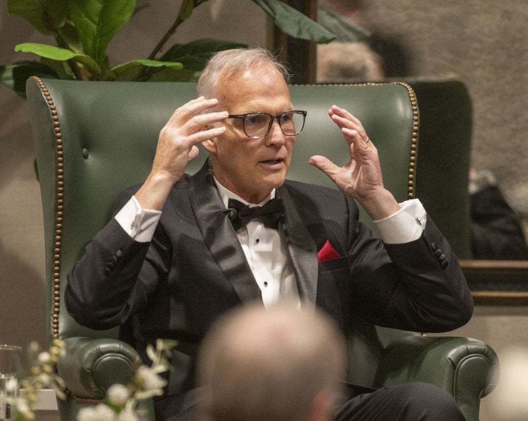 Gentleman sitting onstage talking about something, holding his hands up in an explanatory gesture. (Photo Credit: Wingate Downs)