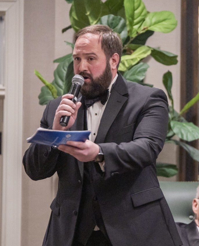 Photo of a man with a beard speaking into a microphone, he appears to be holding que cards. (Photo Credit: Wingate Downs)