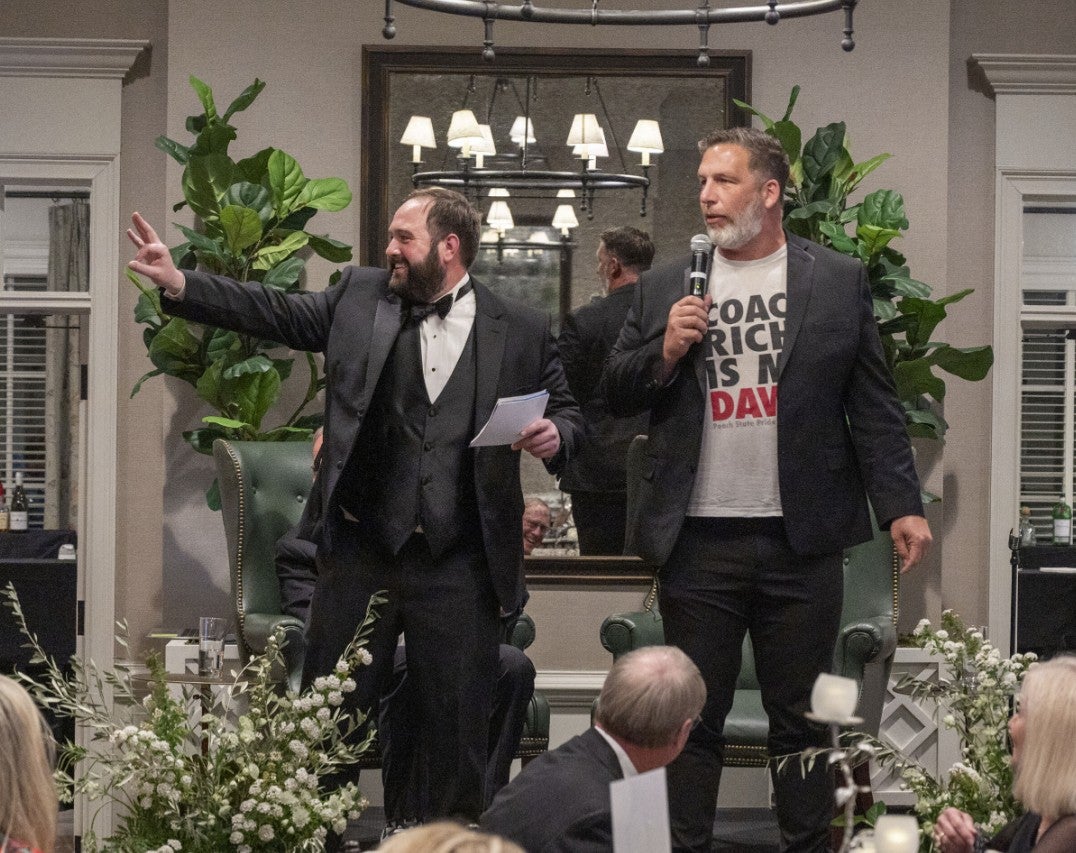Photo of two men on stage holding microphones, speaking to the crowd. (Photo Credit: Wingate Downs)