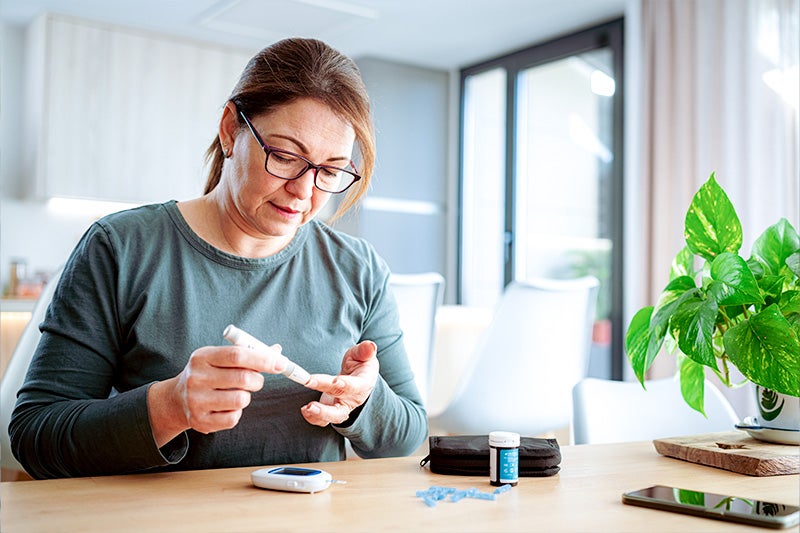 Older woman sitting at a table testing her blood sugar with a diabetic finger prick test.